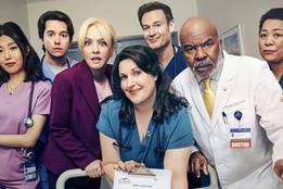 The cast of St. Denis Medical poses in a hospital hallway, dressed in scrubs and lab coats, capturing the show’s comedic hospital vibe.