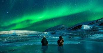 Two people sitting on frozen lake enjoying aurora borealis in Alaska